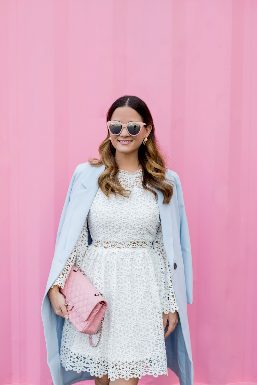 Jennifer Lake Style Charade wearing a white lace bell sleeve dress, long blue coat, Kate Spade Lydia heels, and pink Chanel quilted flap bag at a Chicago pink wall