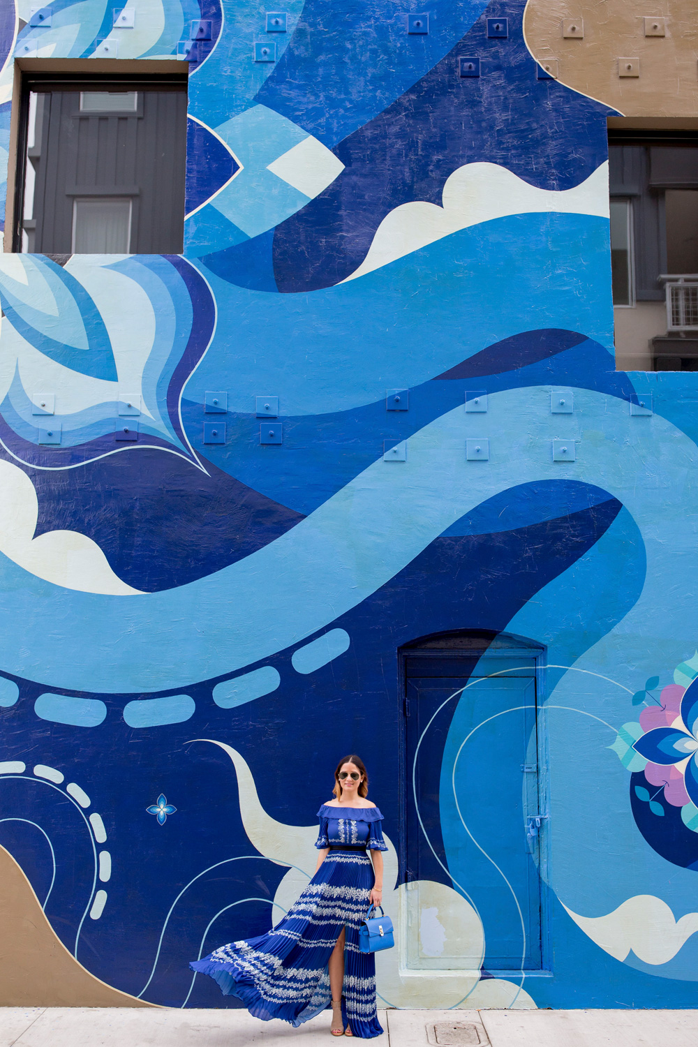 Jennifer Lake Style Charade wearing a blue pleated Self Portrait maxi dress, and blue Henri Bendel bag at a blue swirl mural wall