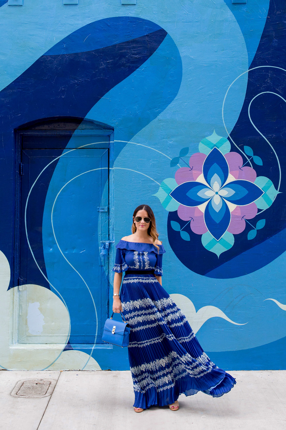 Jennifer Lake Style Charade wearing a blue pleated Self Portrait maxi dress, and blue Henri Bendel bag at a blue swirl mural wall