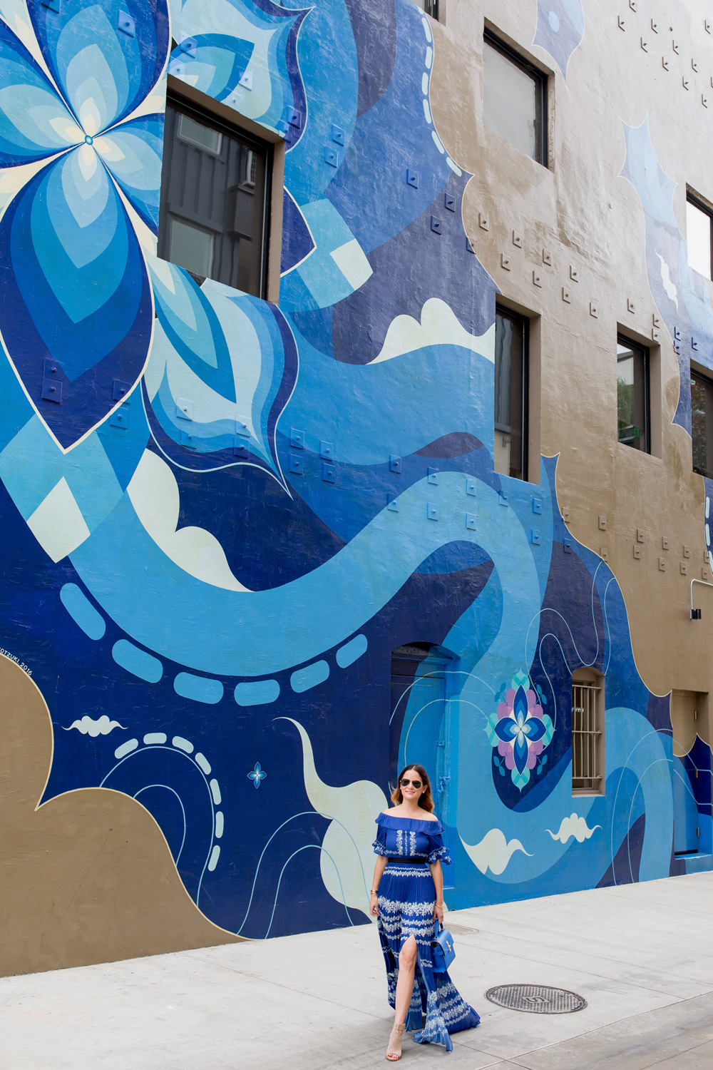 Jennifer Lake Style Charade wearing a blue pleated Self Portrait maxi dress, and blue Henri Bendel bag at a large blue swirl mural wall