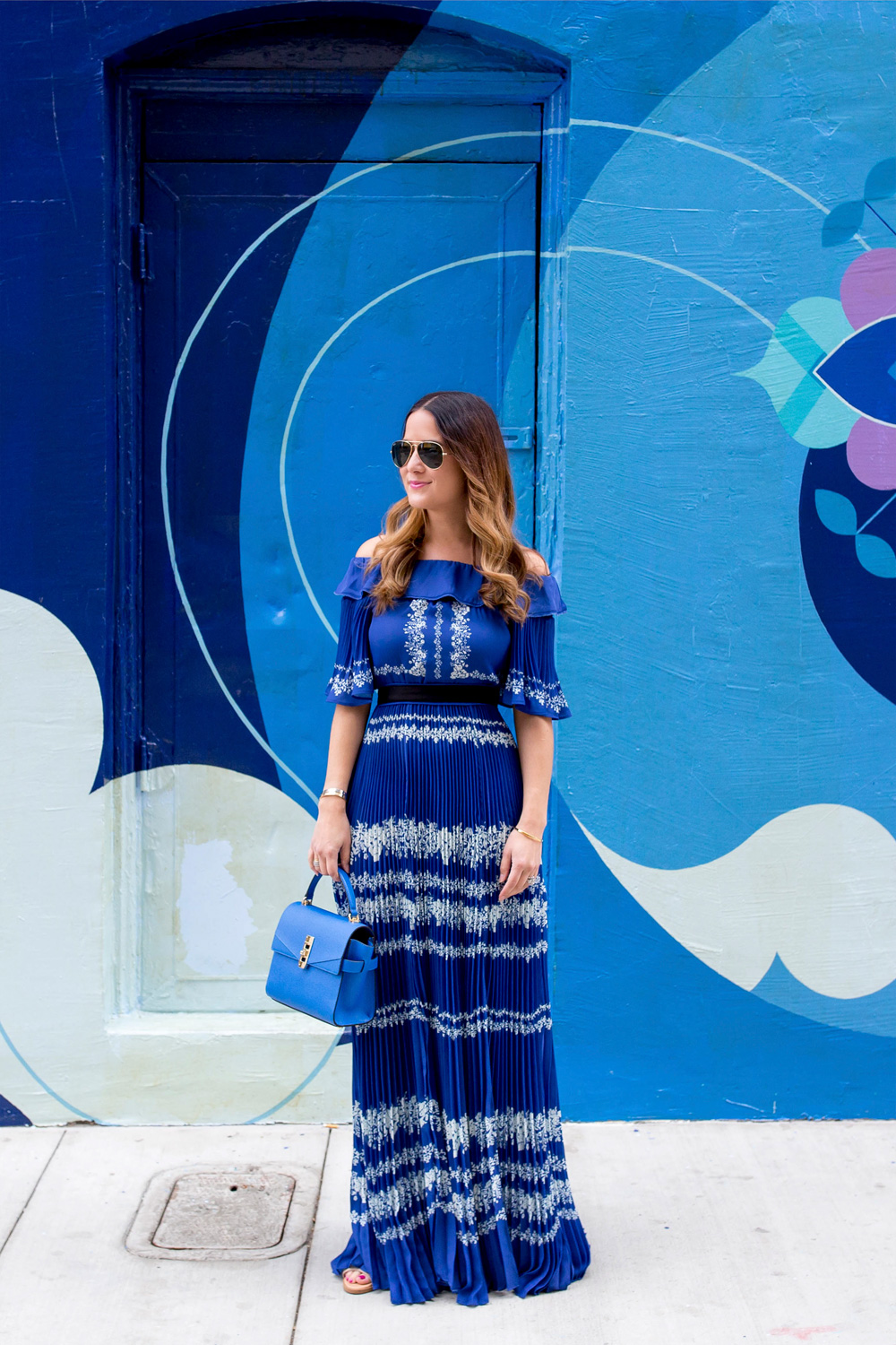 Jennifer Lake Style Charade wearing a blue pleated Self Portrait maxi dress, and blue Henri Bendel bag at a blue swirl mural wall