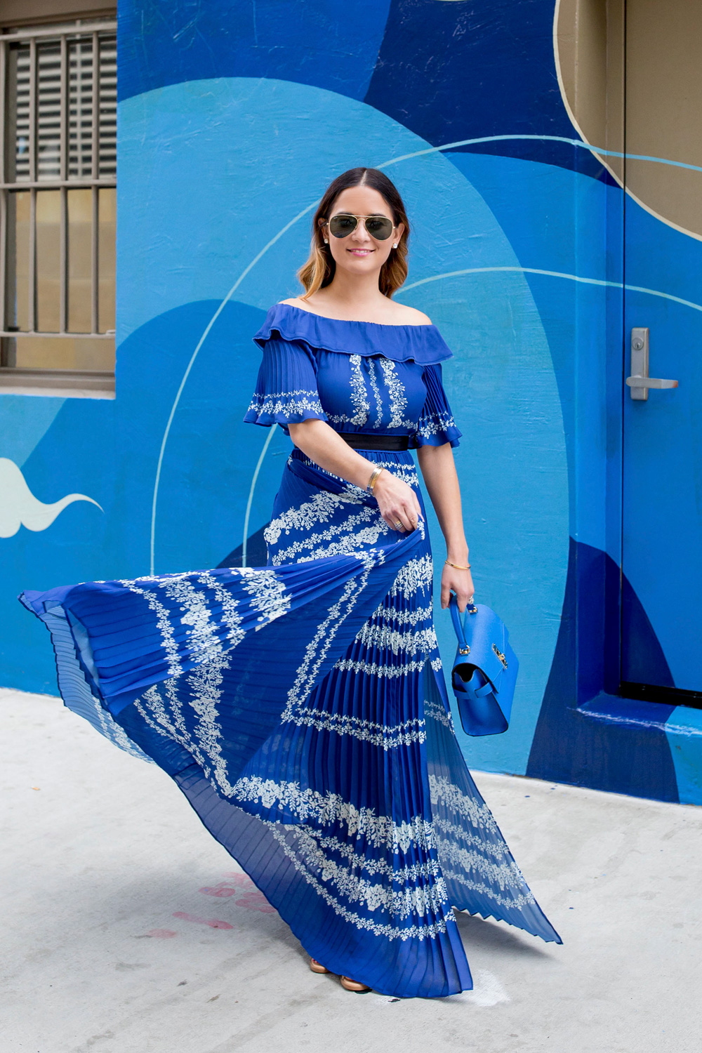 Jennifer Lake Style Charade twirling in a blue pleated Self Portrait maxi dress, and blue Henri Bendel bag at a blue swirl mural wall