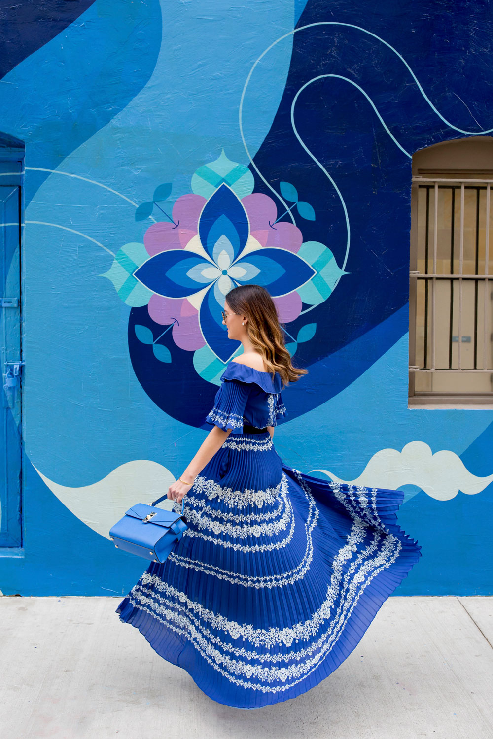Jennifer Lake Style Charade twirling in a blue pleated Self Portrait maxi dress, and blue Henri Bendel bag at a blue swirl mural wall