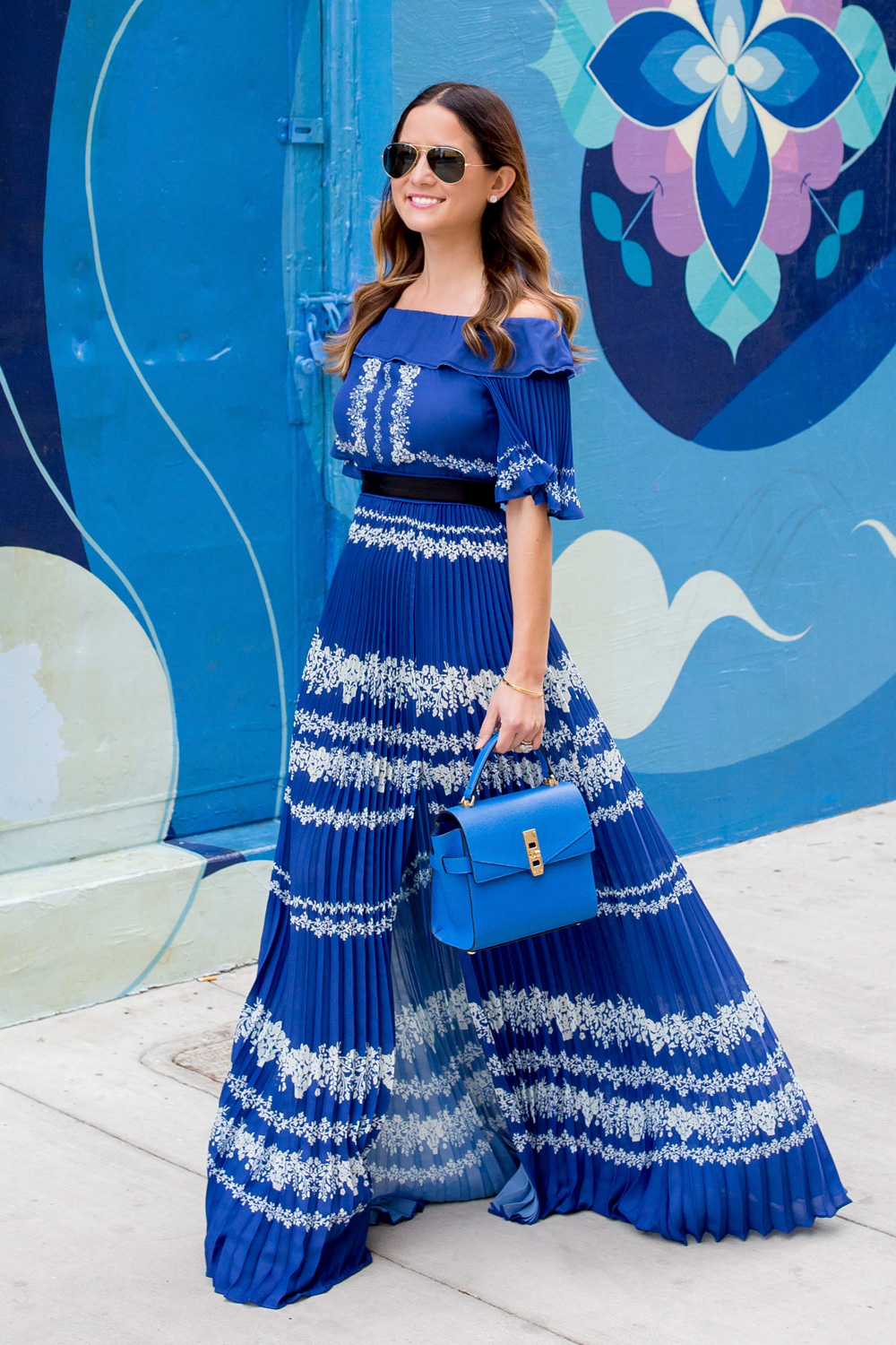 Jennifer Lake Style Charade walking in a blue pleated Self Portrait maxi dress, and blue Henri Bendel bag at a blue street art mural wall