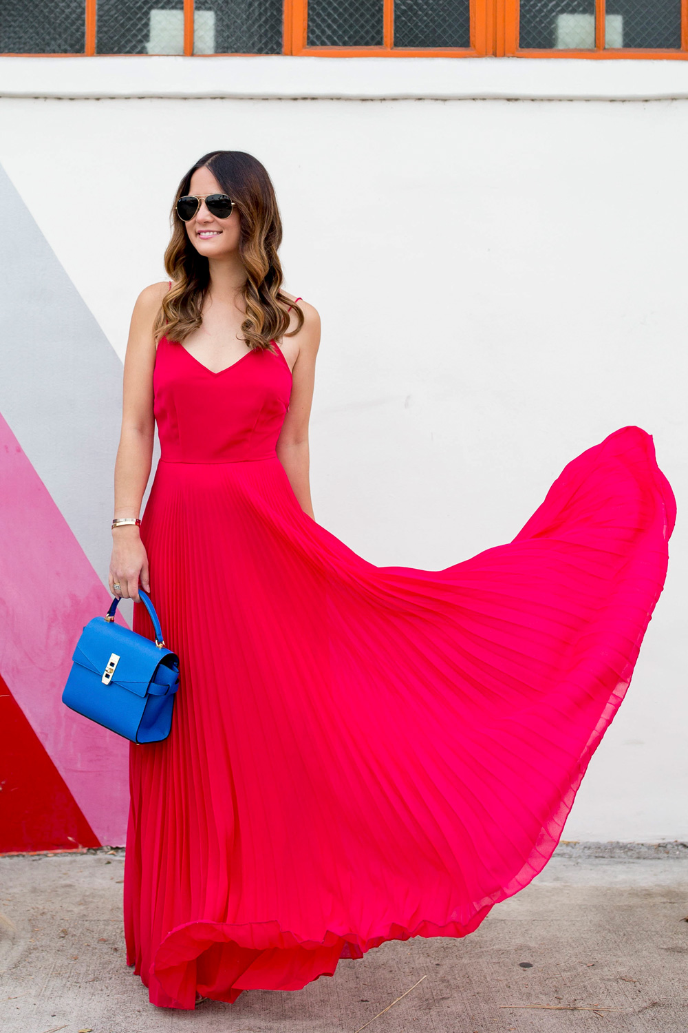 Jennifer Lake Style Charade in a flowing ASOS pink pleated maxi dress, blue Henri Bendel Mini Uptown satchel, at a striped wall in Los Angeles