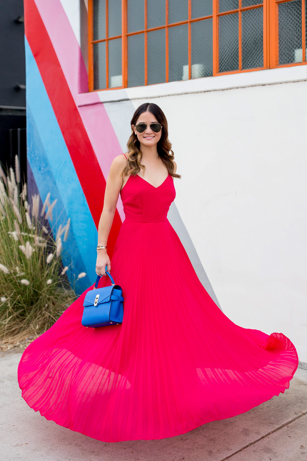 Jennifer Lake Style Charade twirling in an ASOS pink pleated maxi dress, blue Henri Bendel Mini Uptown satchel, at a striped wall in Los Angeles
