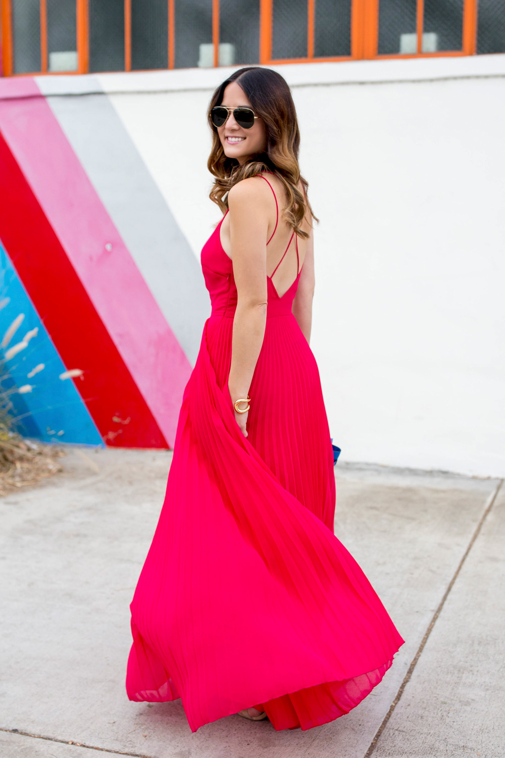 Jennifer Lake Style Charade walking in an ASOS pink pleated maxi dress, blue Henri Bendel Mini Uptown satchel, at a striped wall in Los Angeles