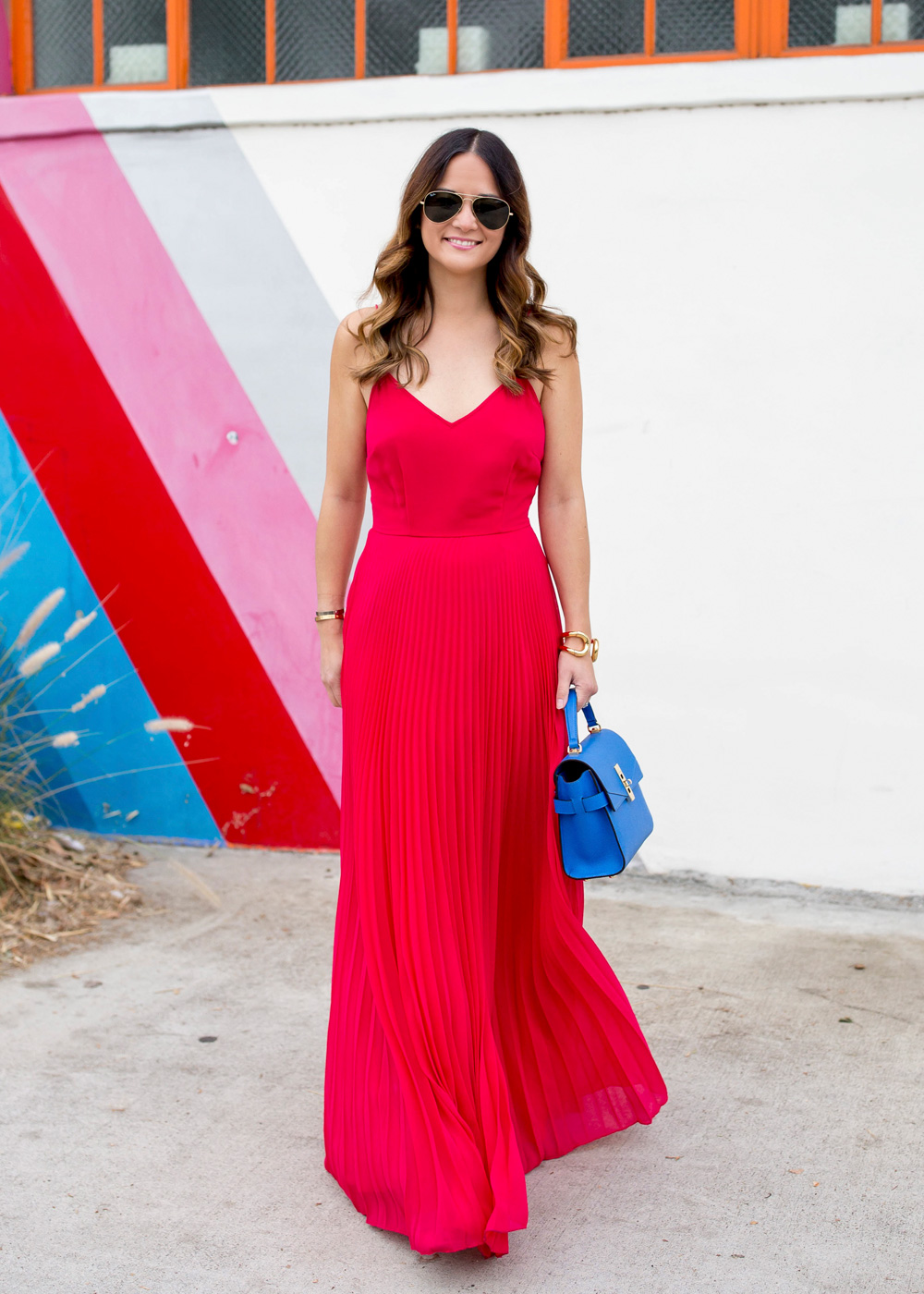Jennifer Lake Style Charade walking in an ASOS pink pleated maxi dress, blue Henri Bendel Mini Uptown satchel, at a striped wall in Los Angeles