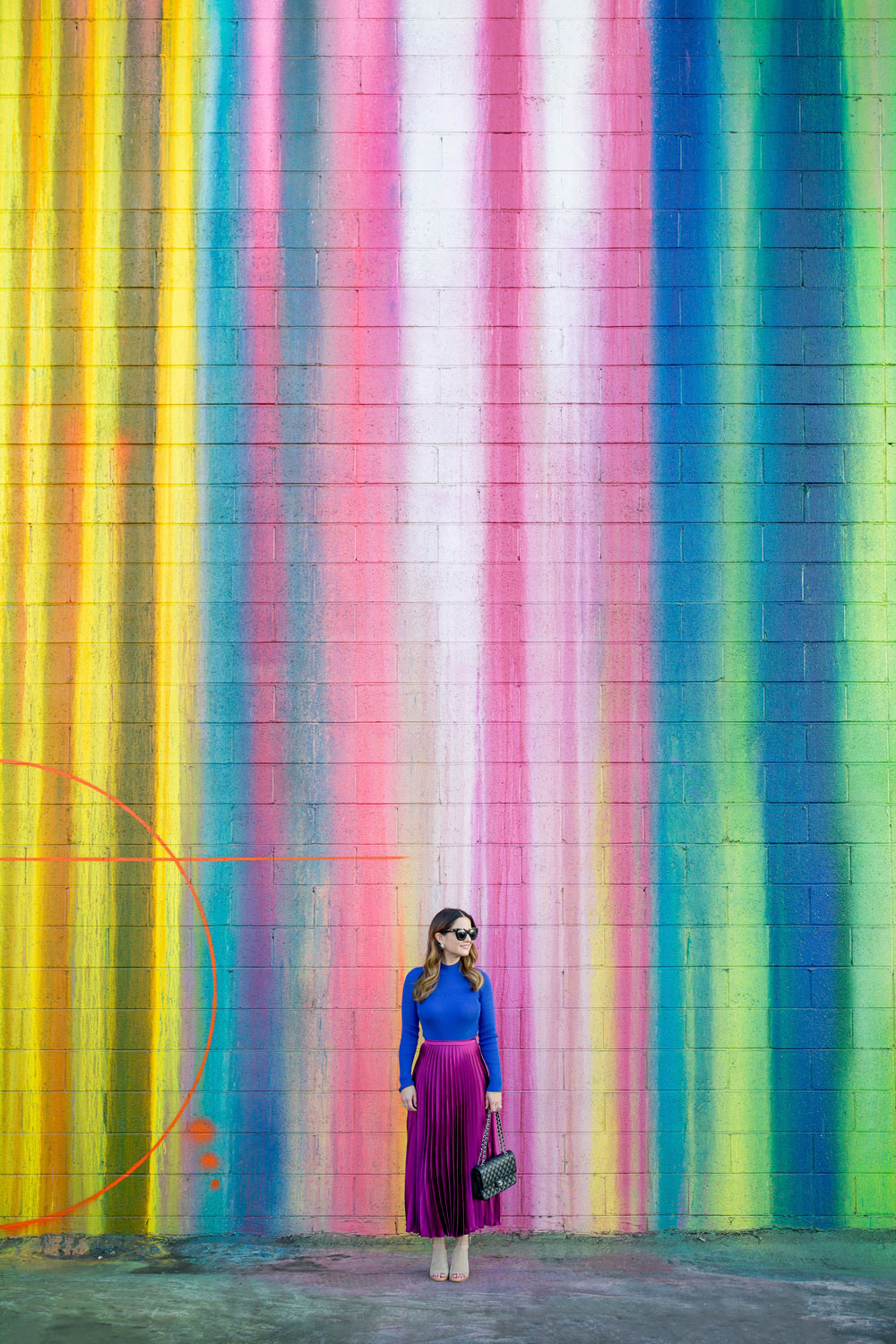 Jennifer Lake Style Charade standing in an ASOS purple pleated midi skirt, cobalt blue sweater bodysuit and Chanel quilted flap bag at a dripping paint wall in Los Angeles