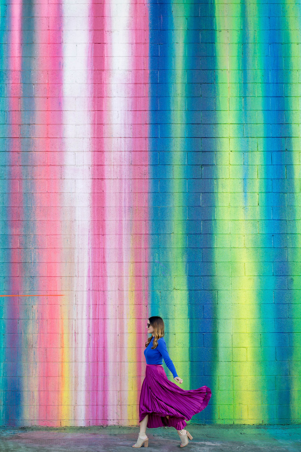 Jennifer Lake Style Charade walking in an ASOS purple pleated midi skirt, cobalt blue sweater bodysuit and Chanel quilted flap bag at a dripping paint wall in Los Angeles