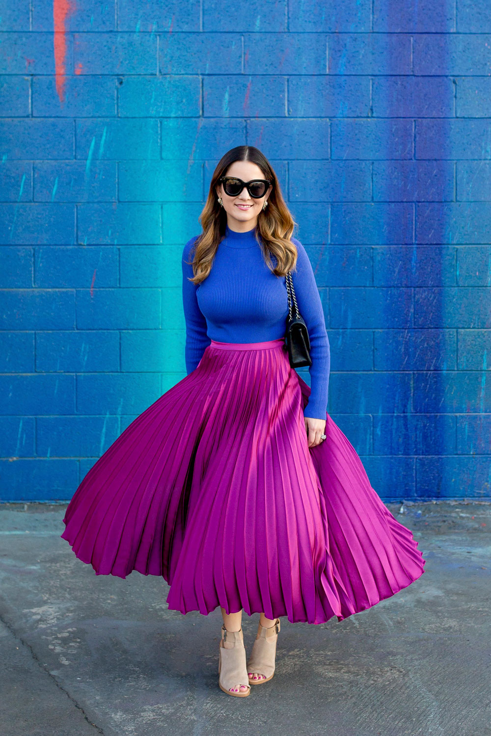 Jennifer Lake Style Charade twirling in an ASOS purple pleated midi skirt, cobalt blue sweater bodysuit and Chanel quilted flap bag at a dripping paint wall in Los Angeles