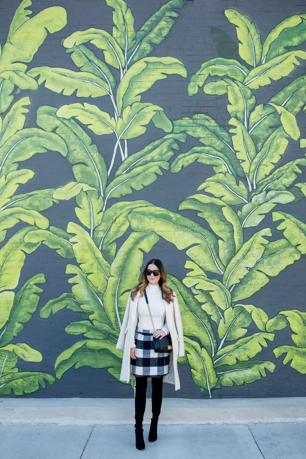 Jennifer Lake Style Charade in an ASOS black white check skirt, ivory coat, Chanel Boy Bag, and Stuart Weitzman Highland boots at a Chicago palm leaf mural