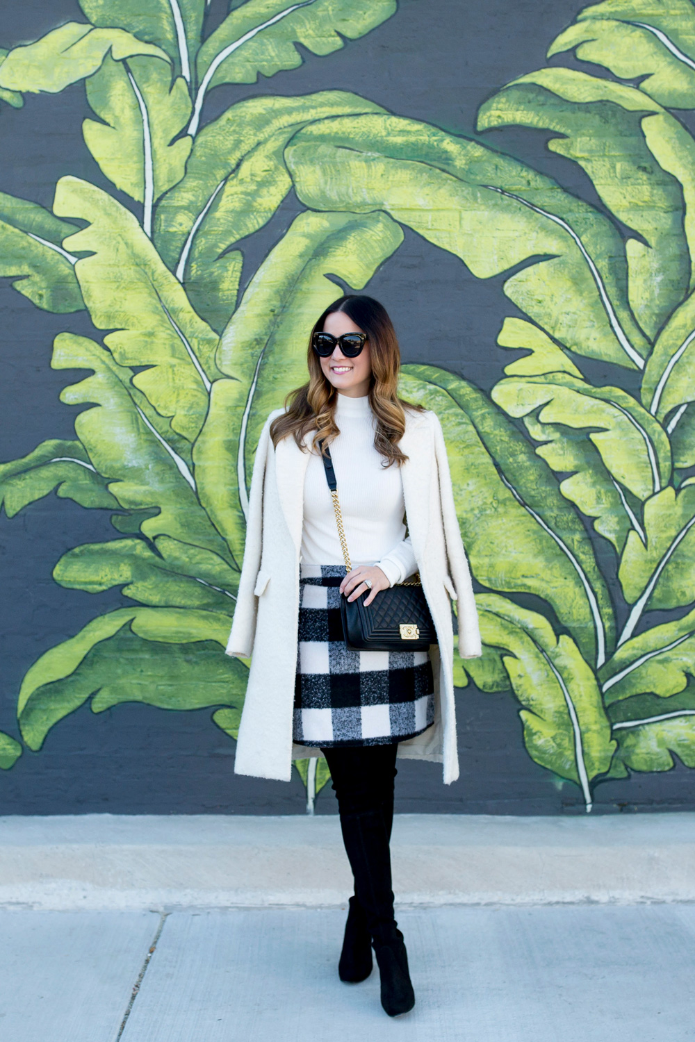 Jennifer Lake Style Charade in an ASOS black white check skirt, ivory coat, Chanel Boy Bag, and Stuart Weitzman Highland boots at a Chicago palm leaf mural