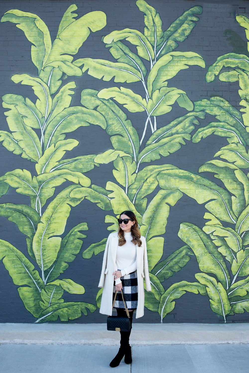 Jennifer Lake Style Charade in an ASOS black white check skirt, ivory coat, Chanel Boy Bag, and Stuart Weitzman Highland boots at a Chicago palm leaf mural