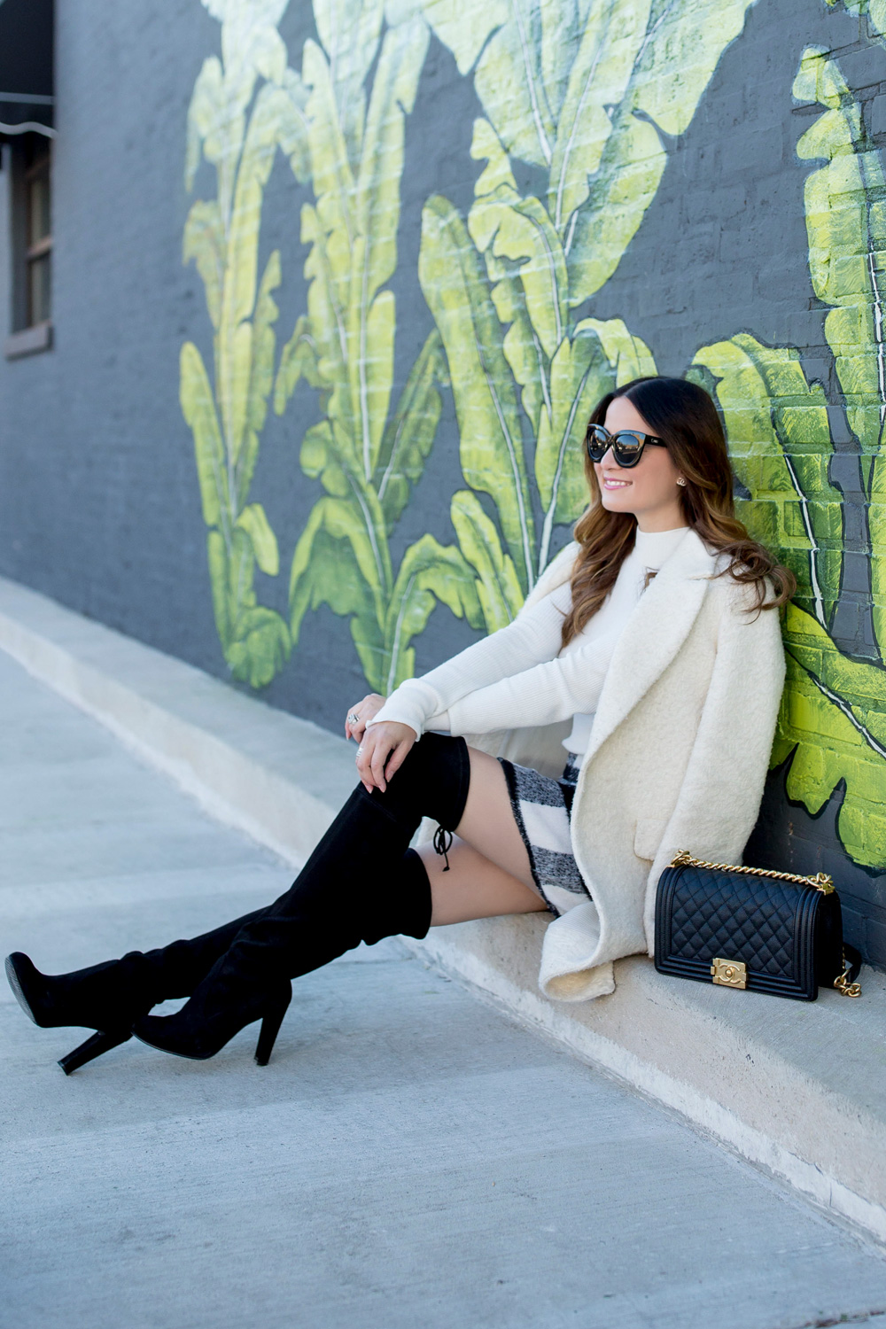 Jennifer Lake Style Charade in an ASOS black white check skirt, ivory coat, Chanel Boy Bag, and Stuart Weitzman Highland boots at a Chicago palm leaf mural