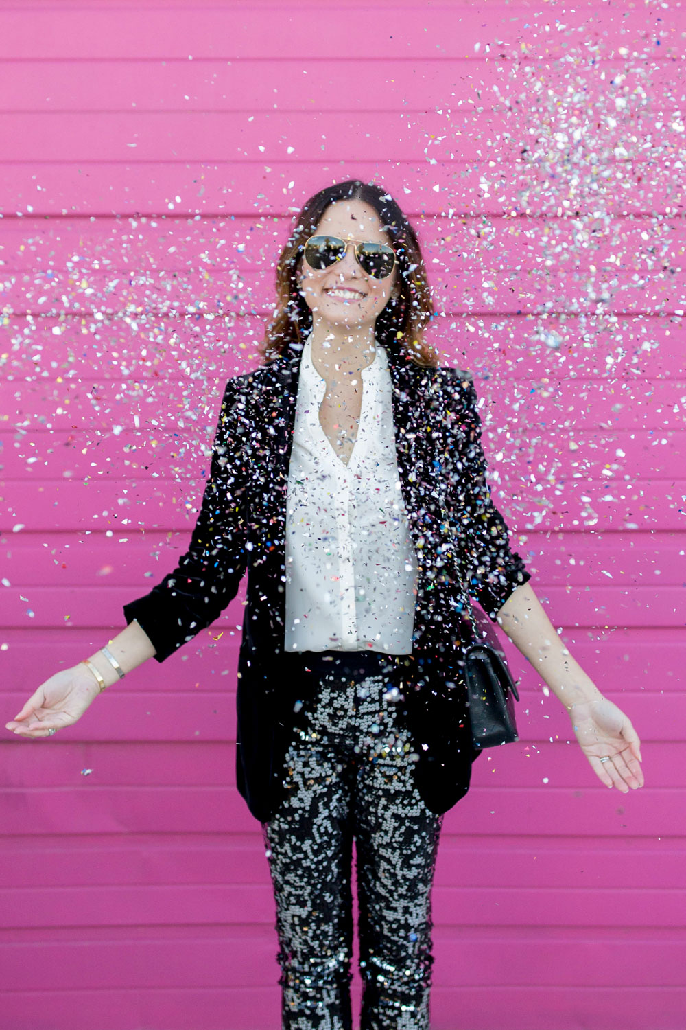 Jennifer Lake Style Charade throwing confetti in Express sequin leggings, black velvet blazer, ivory lace top, and a quilted Chanel flap bag in front of a Chicago pink wall