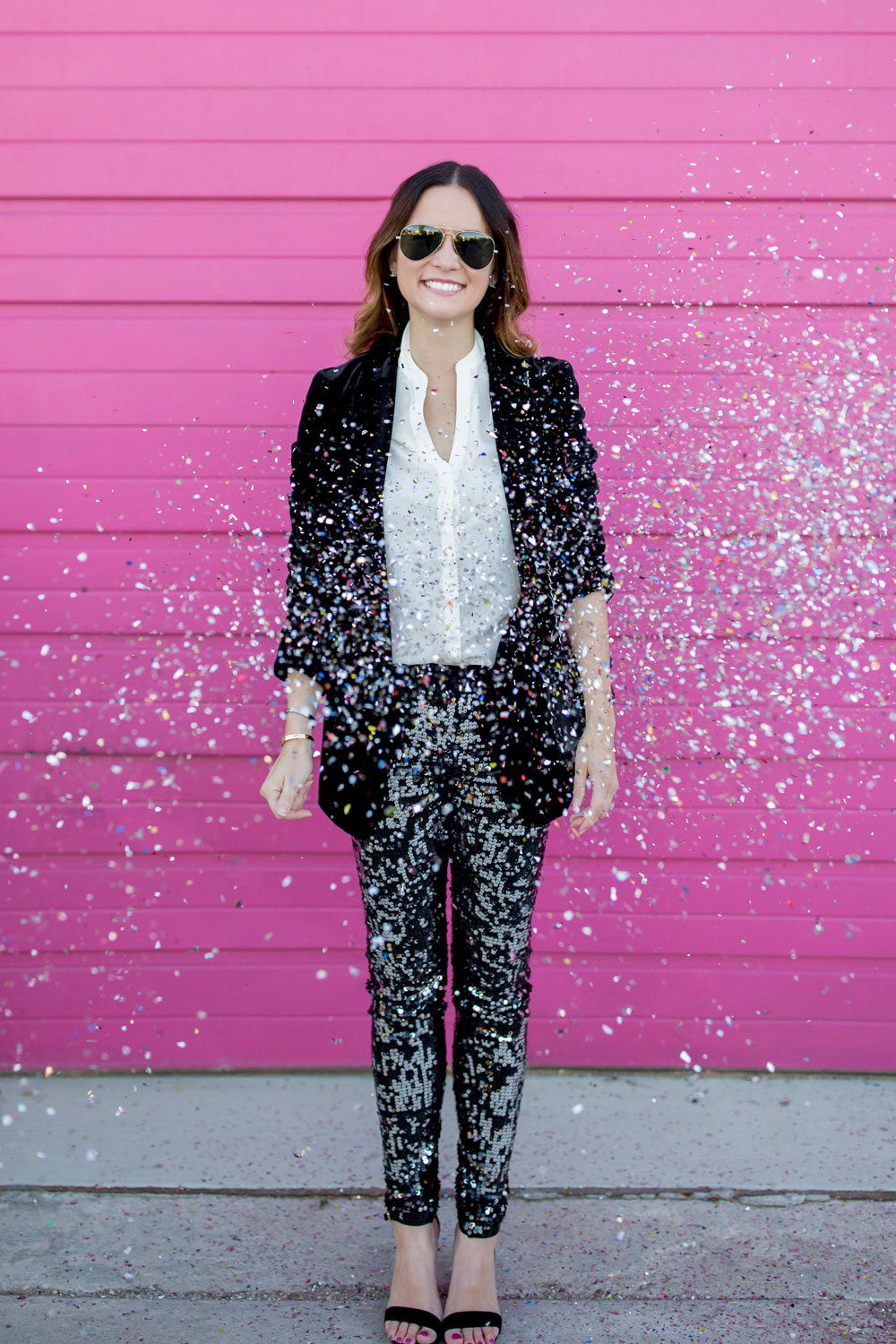 Jennifer Lake Style Charade throwing confetti in Express sequin leggings, black velvet blazer, ivory lace top, and a quilted Chanel flap bag in front of a Chicago pink wall